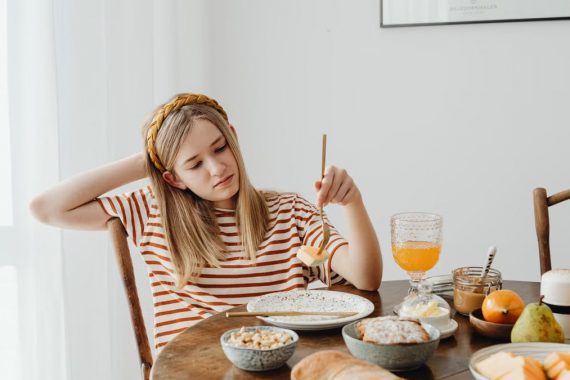 Teen girl in striped shirt at breakfast table with fruits, bread, and juice. Casual morning setting.